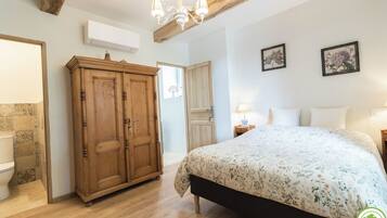 A bedroom with a wooden wardrobe, a bed with floral bedding, a chandelier, and two framed botanical prints on the wall.