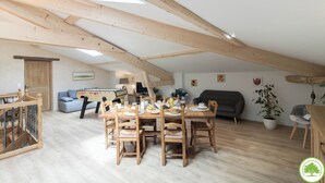 Un loft spacieux avec de hauts plafonds et des poutres apparentes. Il comprend un coin repas avec une table et des chaises en bois, un canapé et un petit espace de jeux avec une table à jouets.