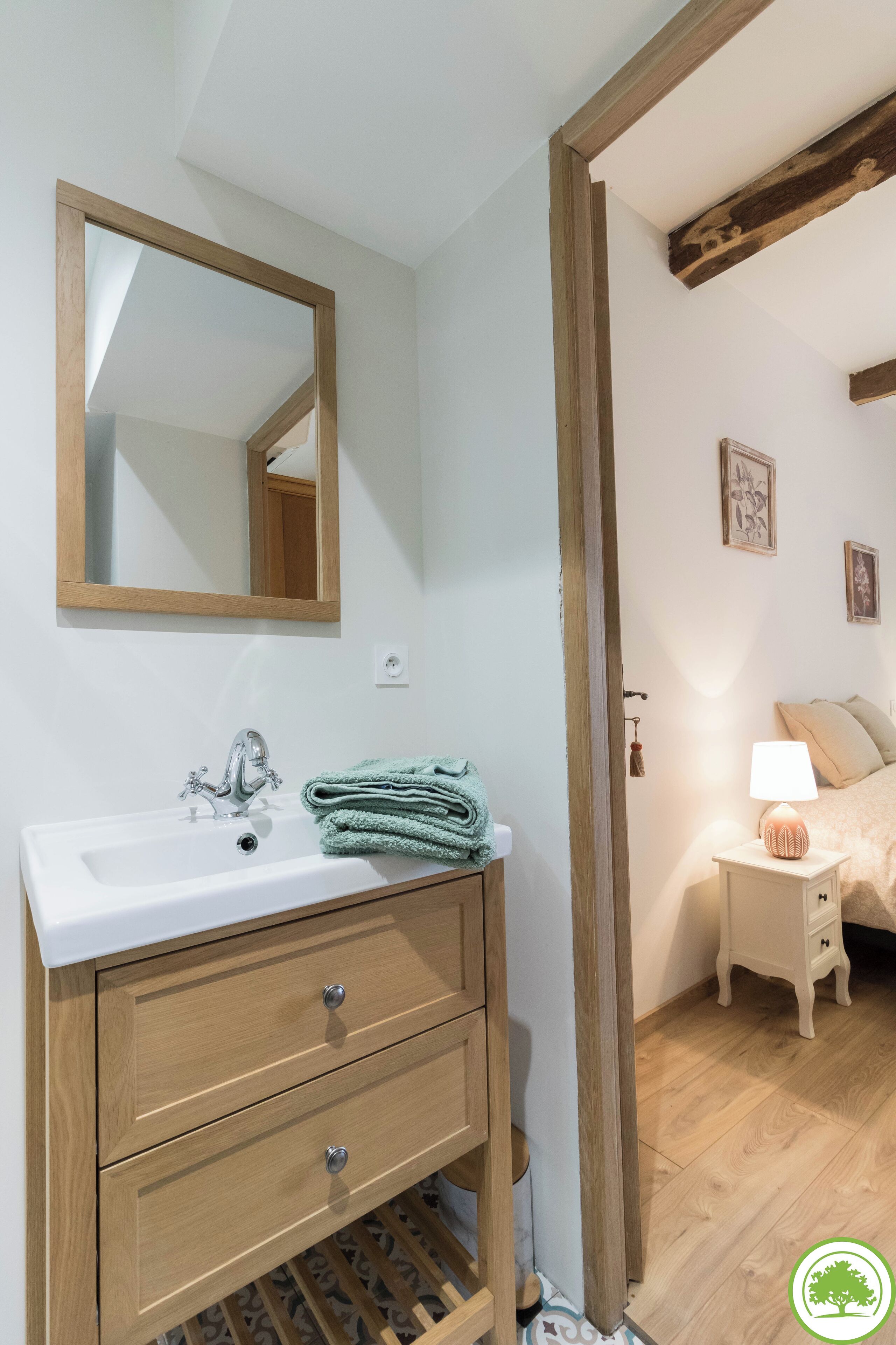 A bathroom with a white sink, wooden vanity, and a mirror. There is a towel on the vanity and a bedside table with a lamp next to a bed.