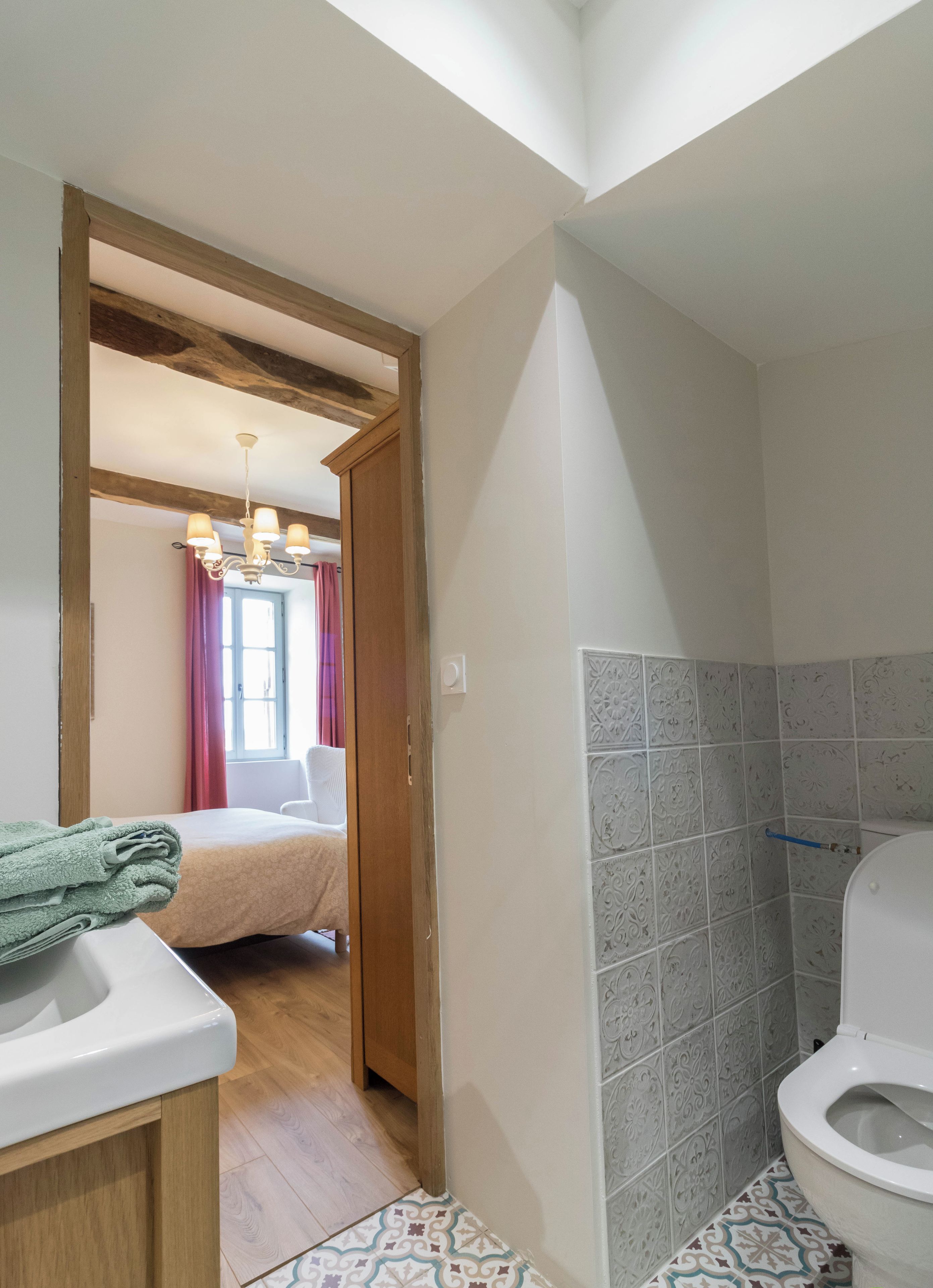 Een badkamer met toilet, wastafel en douchecabine. Een houten deur leidt naar een slaapkamer met een bed en een kroonluchter.