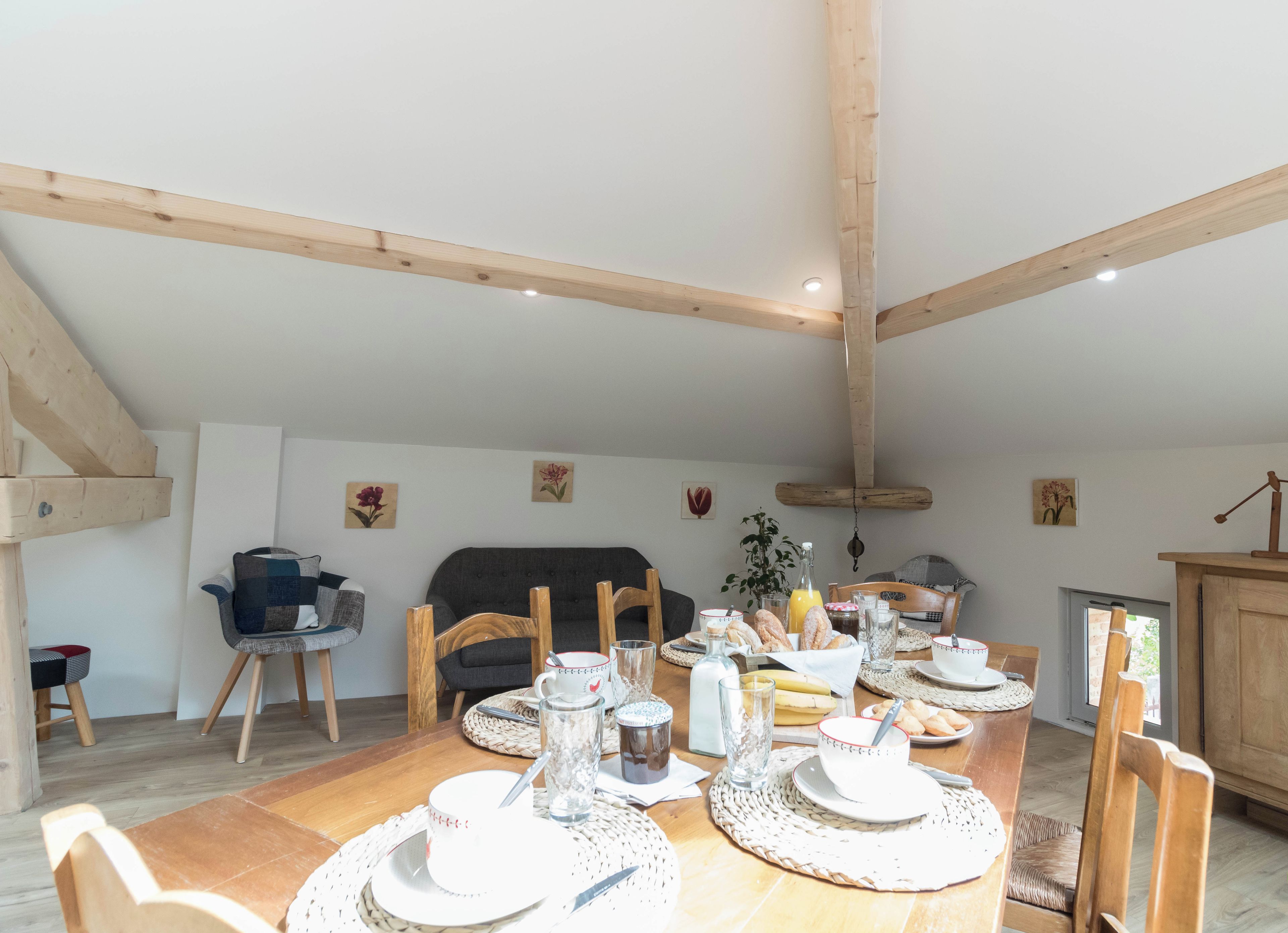 A dining area with a wooden table set for breakfast, surrounded by chairs, with a sofa and a cabinet in the background.