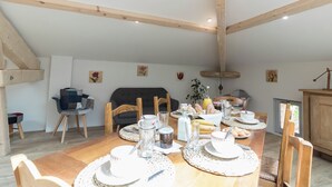 A dining area with a wooden table set for breakfast, surrounded by chairs, with a sofa and a cabinet in the background. - La Clef des Champs (Sajas)