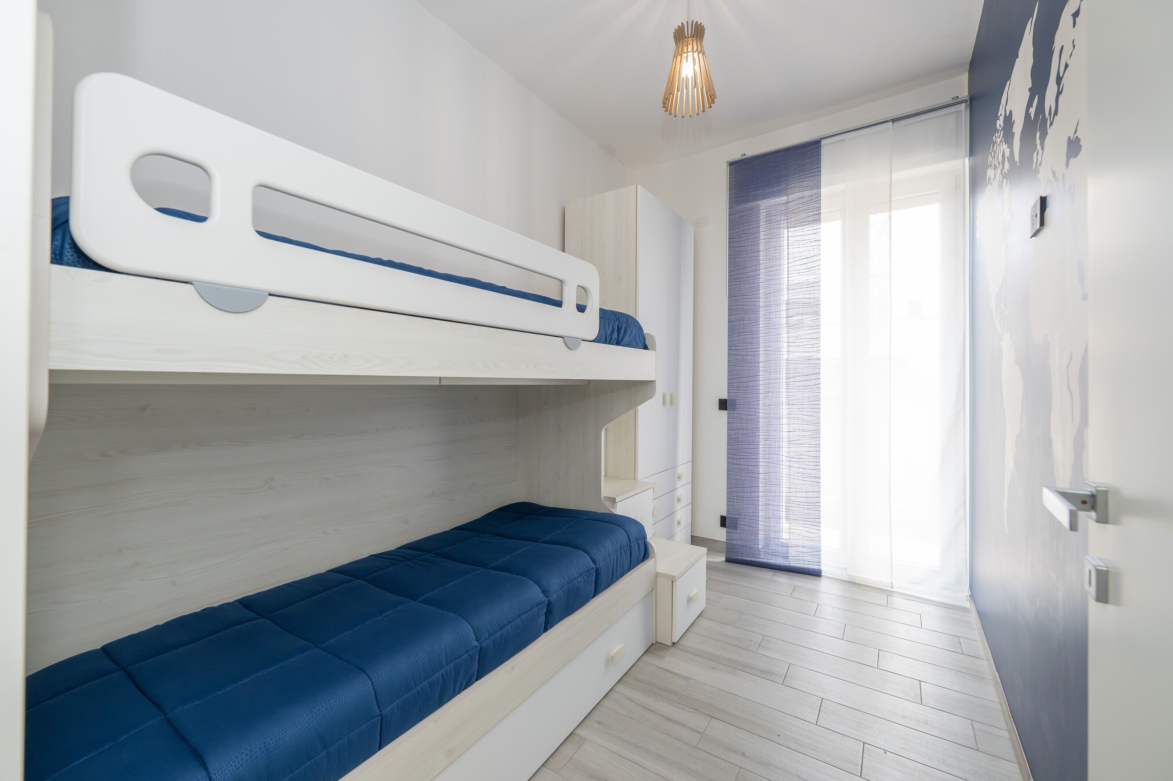 Een kamer met een stapelbed, een klein bureau en een raam met zonwering.