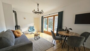 A modern living room with a sofa, a coffee table, and a dining table with chairs. There is a fireplace, a large window with curtains, and a chandelier. - Almrose Zauber, vacation apartment in the mountains near the ski slopes (Oberau)