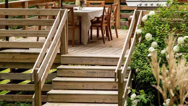 Eine Holzterrasse mit Tisch, Stühlen, einem Grill und einem Sonnenschirm.