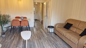 A living room with a beige sofa, a dining table with orange chairs, and a white chair. - NightRest Homes Orchid2 NEC Free Parking  2Toilets (West Midlands)