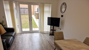 A room with a wooden floor, a black leather sofa, a wooden dining table with chairs, a flat-screen TV, and a clock on the wall. - NightRest Homes Orchid NEC Free Parking  2 Toilets (West Midlands)