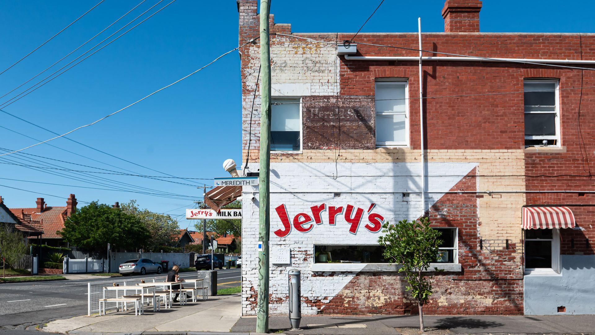 A building with a prominent red sign reading 'Jerry's' on a white wall, a street sign above it, and a small tree in front.