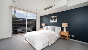 A bedroom with a large sliding glass door leading to a balcony, a bed with white bedding, two bedside tables with lamps, and a dark-colored accent wall with a framed picture.