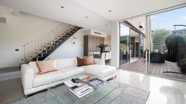 A modern living room with a white sectional sofa, a glass coffee table with magazines, a staircase, and a sliding glass door leading to an outdoor patio.