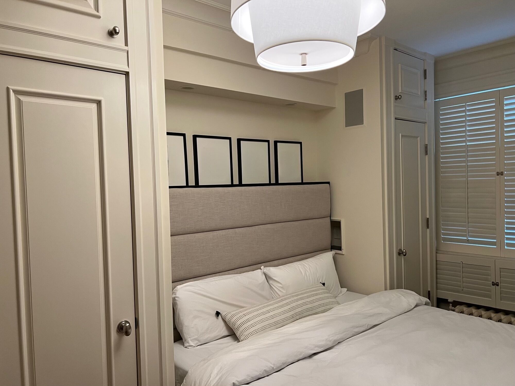 Un dormitorio con cama, cabecera con cuadros enmarcados, una puerta y una ventana con persianas.