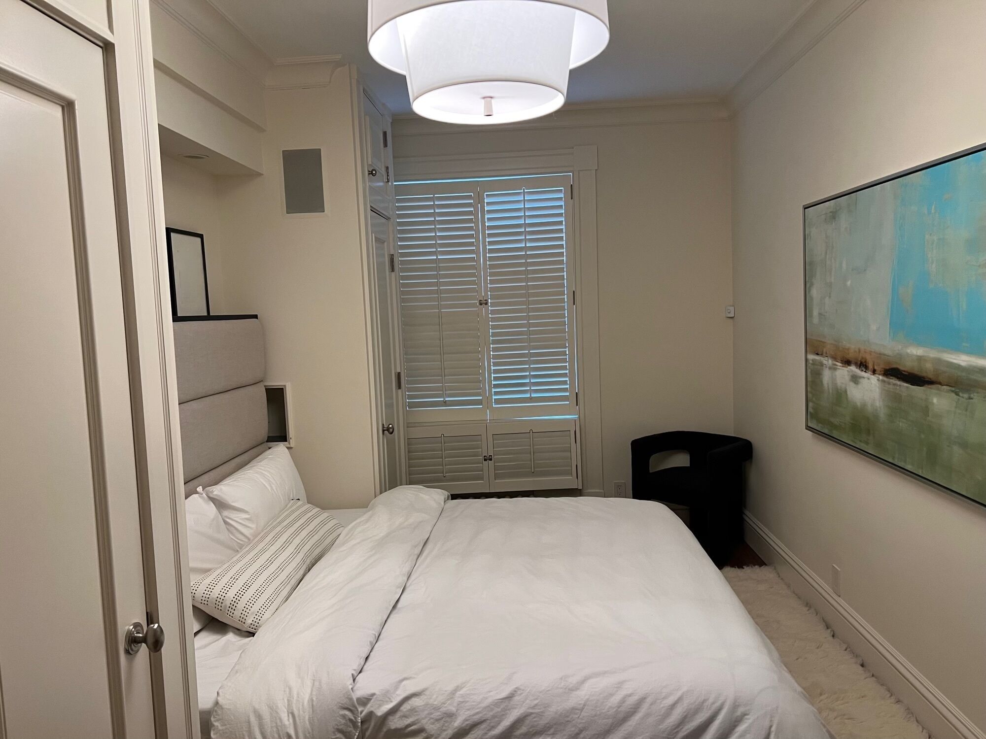 A bedroom with a bed, a door, a window with blinds, a ceiling light, and a painting on the wall.