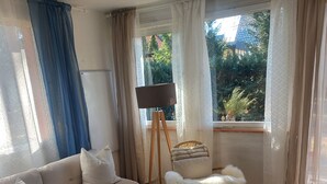 A living room with a sofa, a coffee table, a floor lamp, a white furry chair, and a view of greenery outside. - NEW "Natur.lich Müritz" (Röbel / Müritz)