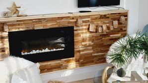 A living room with a fireplace, a flat-screen TV mounted above it, and a round glass table with a plant. - NEW "Natur.lich Müritz" (Röbel / Müritz)