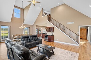 A spacious living room with a black leather sectional sofa, a wooden coffee table, and a dining area with a wooden table and chairs. A kitchen with wooden cabinets and a staircase leading to an upper level is visible. - Hot Tub + Fire Pit | Private Country Escape (Pacific)