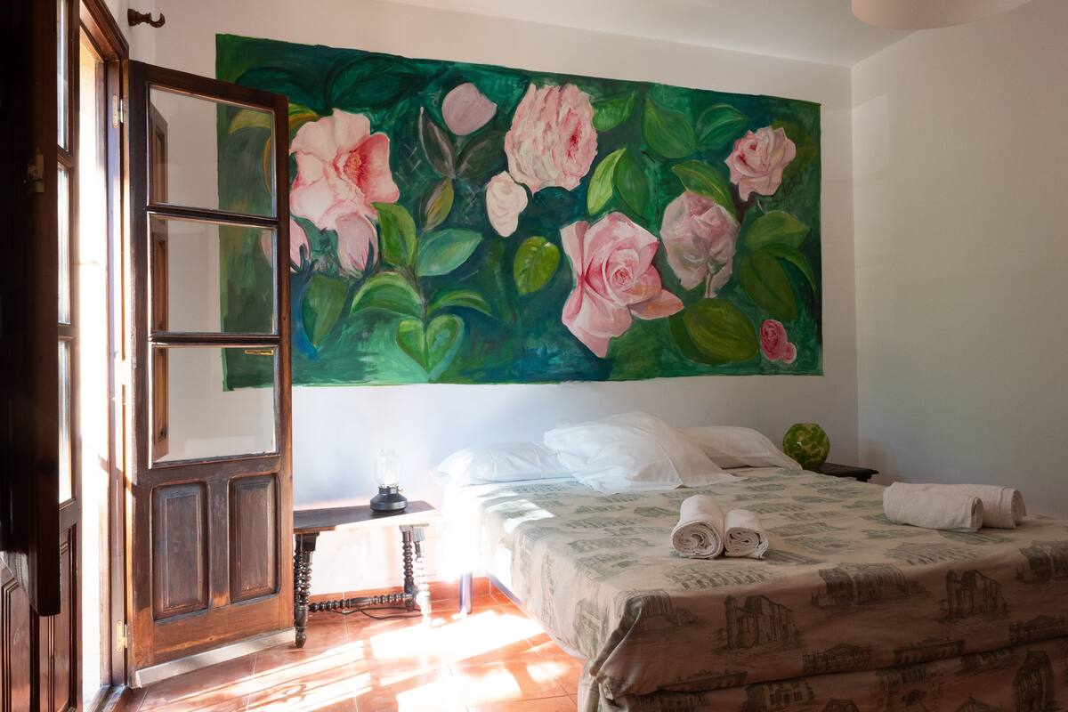A hotel room with a bed, a painting of pink roses on the wall, a wooden door, and a nightstand with a lamp.
