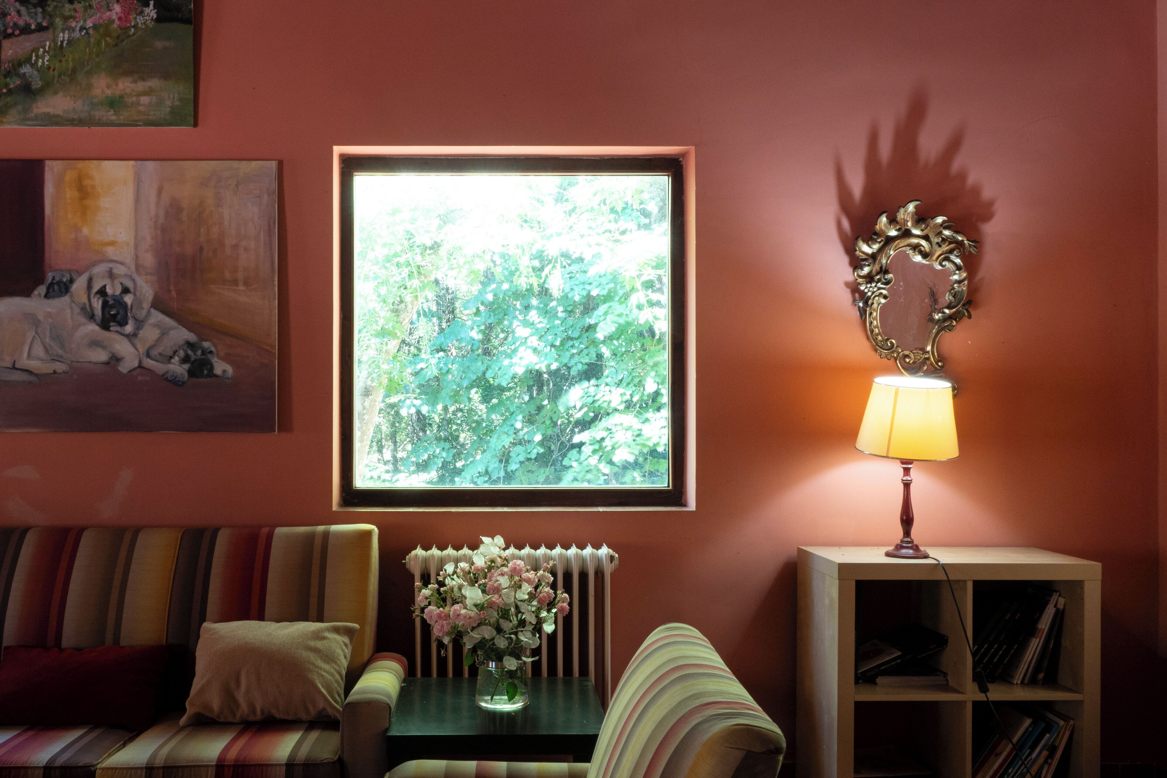 Una sala de estar con sofá, una mesita con un florero, una estantería, un cuadro enmarcado de un perro y una ventana con vistas a árboles.