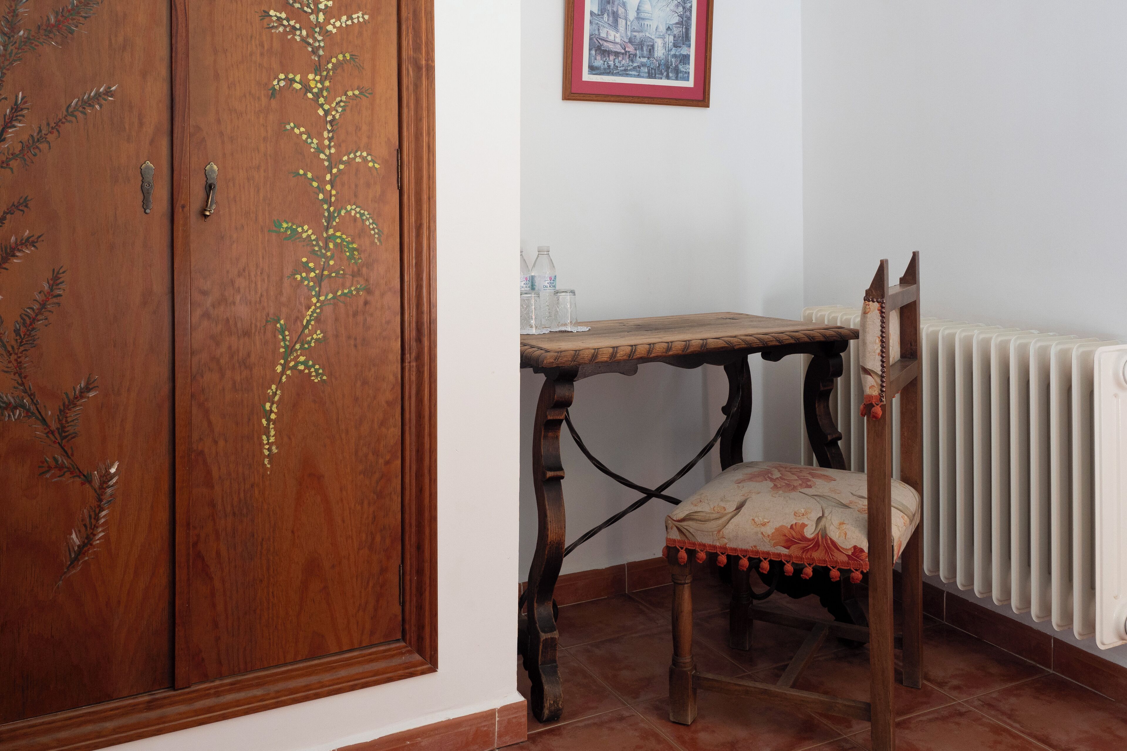Una puerta de madera con adornos florales, una silla de madera con cojín floral, una mesita de madera y un radiador.