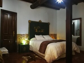 Quarto com porta de madeira, parede de destaque em tijolo, cabeceira verde, cama com roupa de cama branca, mesa de cabeceira com abajur e tapete de couro de vaca.