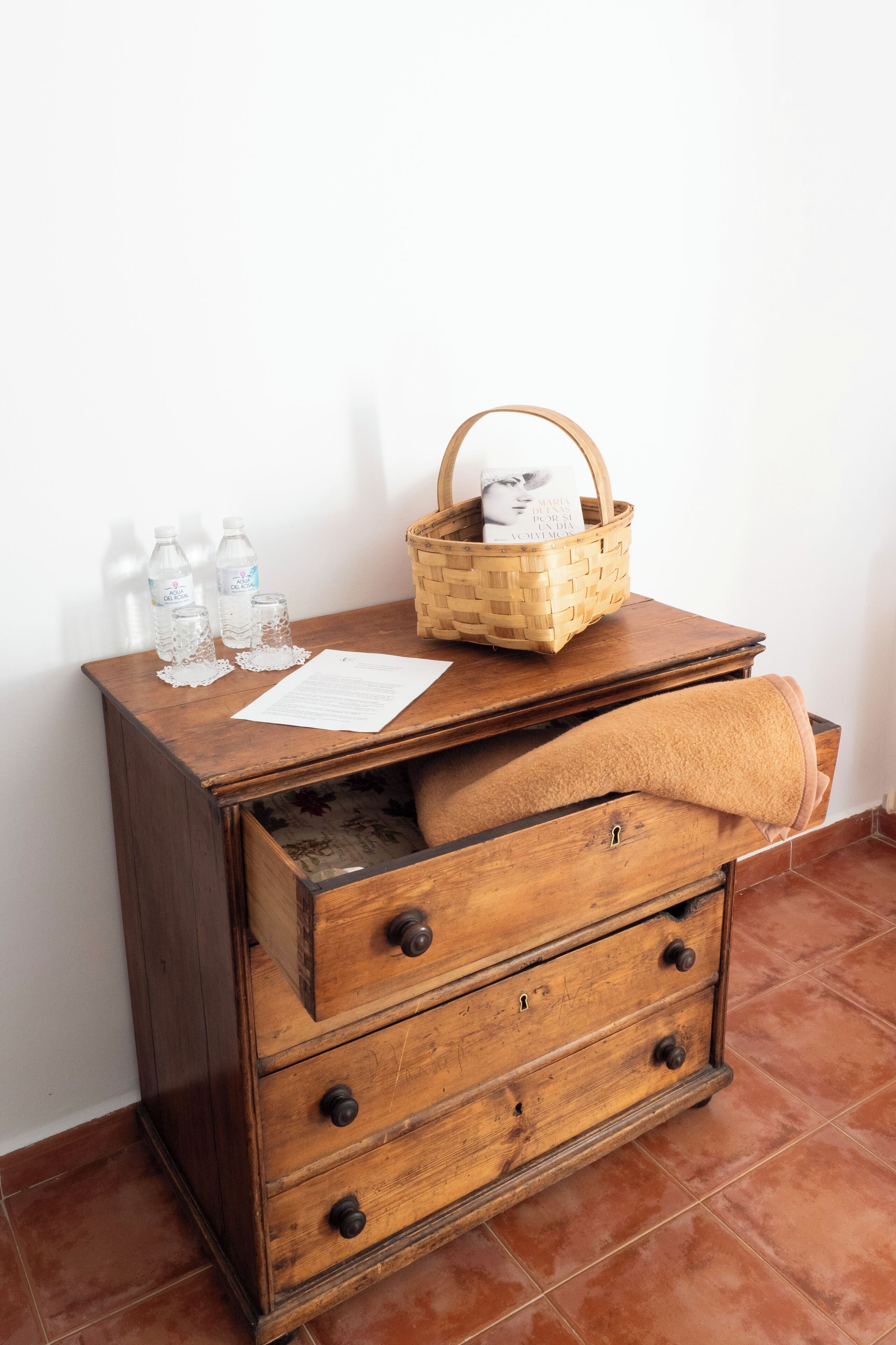 Une commode en bois surmontée d’un panier en osier, une serviette pliée et des bouteilles d’eau, le tout posé sur une table en bois.