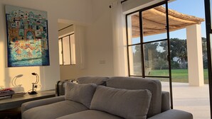 Ett modernt vardagsrum med en grå soffa, en målning som föreställer ett kustlandskap och ett stort fönster med utsikt över en trädgård och en träpergola.