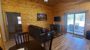 Interior de uma cabana de madeira com sofá, televisão, lareira e uma porta de vidro deslizante para área externa.