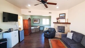 A living room with a fireplace, a television, a ceiling fan, and a kitchen area with a refrigerator and a sink. - Pine Creek Cabins - Cabin 5 (L2) (Pine)