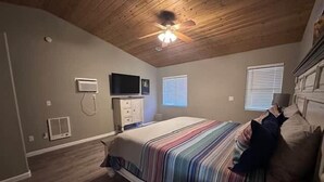 Una camera da letto con soffitto in legno, un letto con biancheria a righe, una televisione a parete e una finestra con persiane.