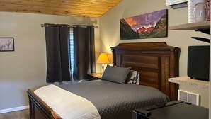 A bedroom with a wooden ceiling, a bed with a headboard, a nightstand with a lamp, a painting on the wall, and an air conditioning unit.