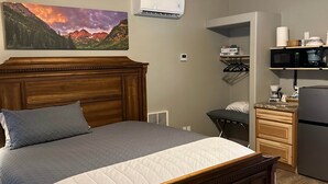 A bedroom with a wooden ceiling, a bed with a headboard, a mounted air conditioner, a desk with a computer, and a kitchen area with a microwave.