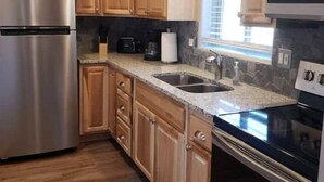 A compact kitchen with wooden cabinets, a stainless steel refrigerator, a black stove, and a window with blinds. - Pine Creek Cabins - Cabin 1 (Pine)