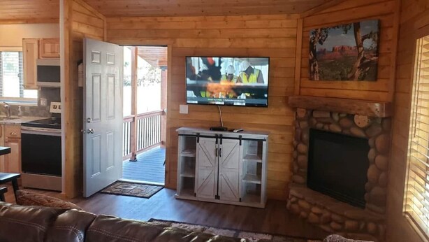 A wooden cabin interior with a stone fireplace, a television mounted above it, and a kitchen area visible through an open door. - Pine Creek Cabins - Cabin 1 (Pine)