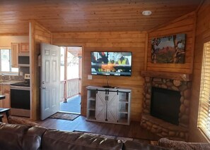 Interior de una cabaña de madera con chimenea de piedra, un televisor montado encima y una cocina visible a través de una puerta abierta.