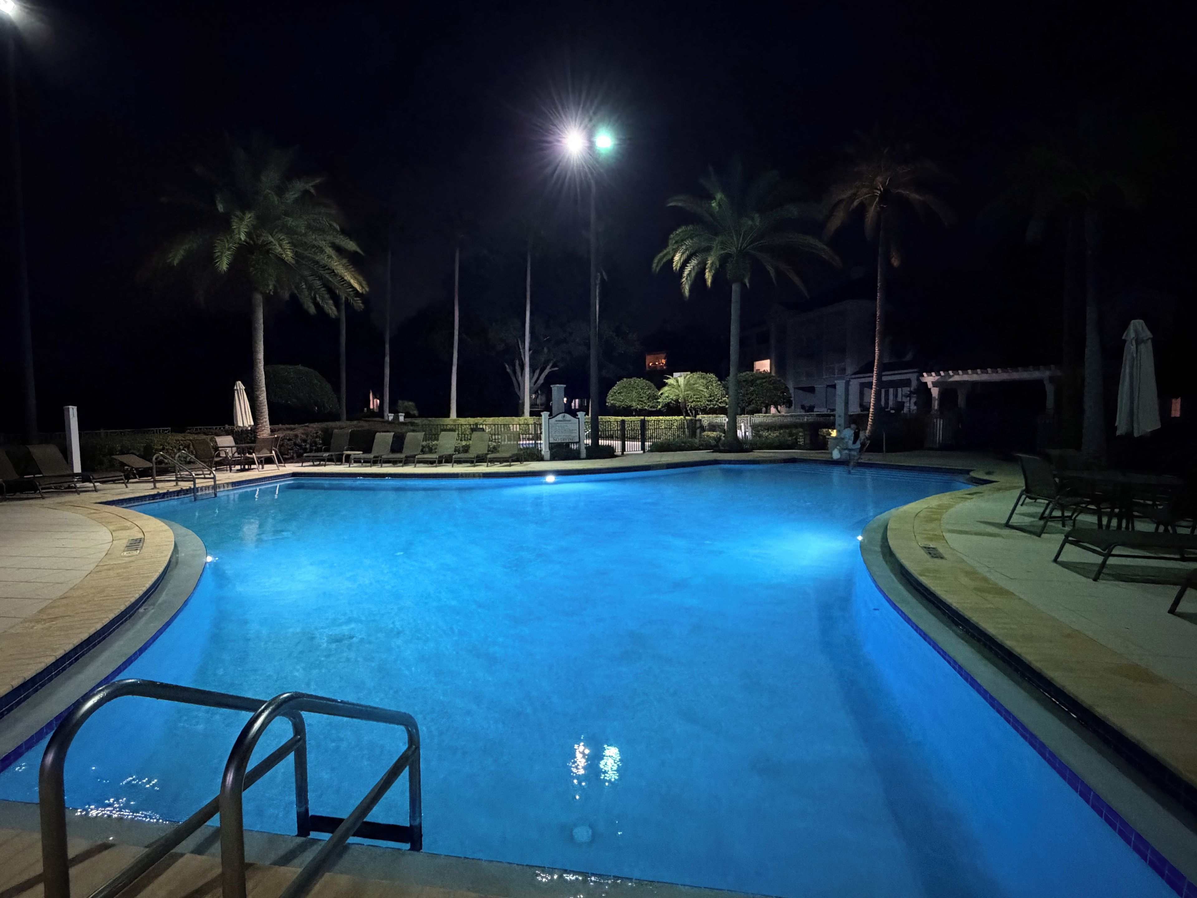 A nighttime swimming pool with a clear water surface, surrounded by lounge chairs and palm trees, under bright artificial lighting.