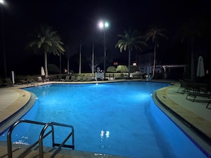Une piscine nocturne à l’eau claire, entourée de chaises longues et de palmiers, illuminée par un éclairage artificiel vif.