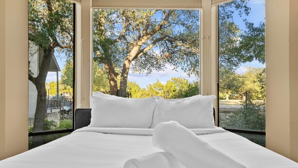A hotel room with a large bed, white linens, and a view of trees and a house through the window. - Lakeway Hill Country Home | Grill Deck, Tennis Courts + Trails (Lakeway)