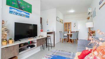 A modern living room with a flat-screen TV, a dining area with a wooden table and chairs, and a grey sofa.