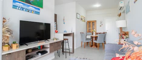 Una sala de estar moderna con un televisor de pantalla plana, un comedor con mesa y sillas de madera, y un sofá gris.