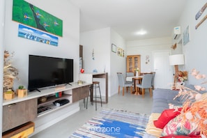 A modern living room with a flat-screen TV, a dining area with a wooden table and chairs, and a grey sofa. - Apart w/ Barbecue in Campeche Clc0104 (Florianópolis)