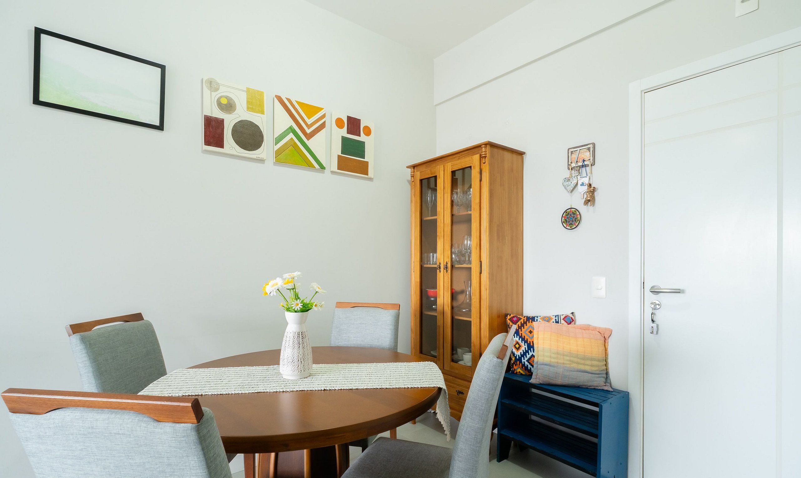 Un comedor con una mesa redonda de madera, cuatro sillas grises tapizadas, un jarrón blanco con flores y un aparador con puertas de cristal.