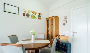 A dining area with a round wooden table, four grey upholstered chairs, a white vase with flowers, and a cabinet with glass doors. - Apart w/ Barbecue in Campeche Clc0104 (Florianópolis)
