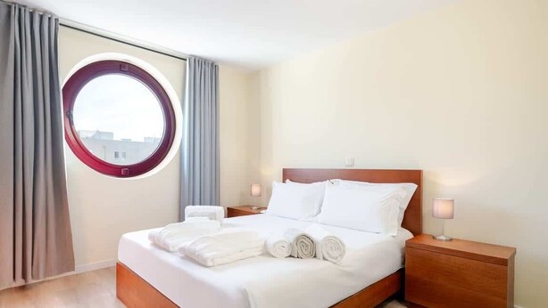 Hotellihuone, jossa on sänky, pyöreä ikkuna, verhot, yöpöytä ja lamppu.