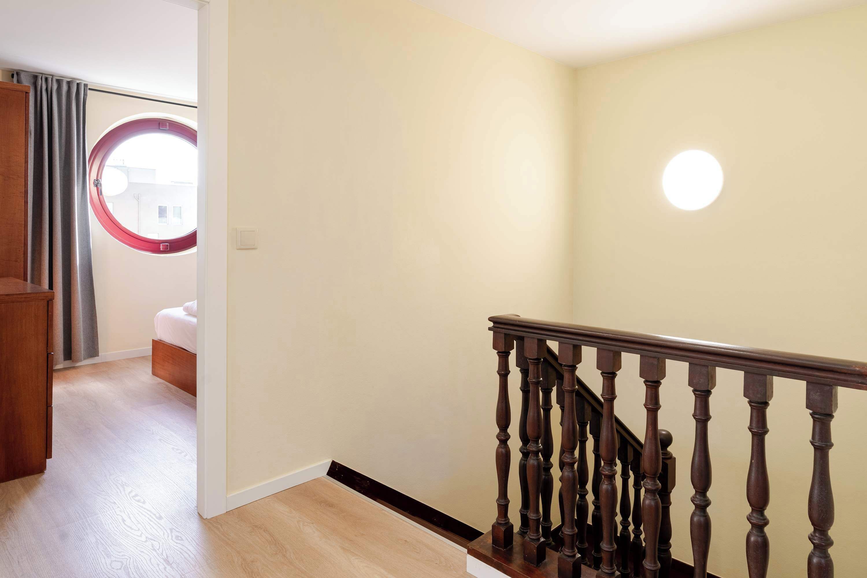 A room with a wooden staircase, a bed, a wardrobe, and a view through a window with curtains.