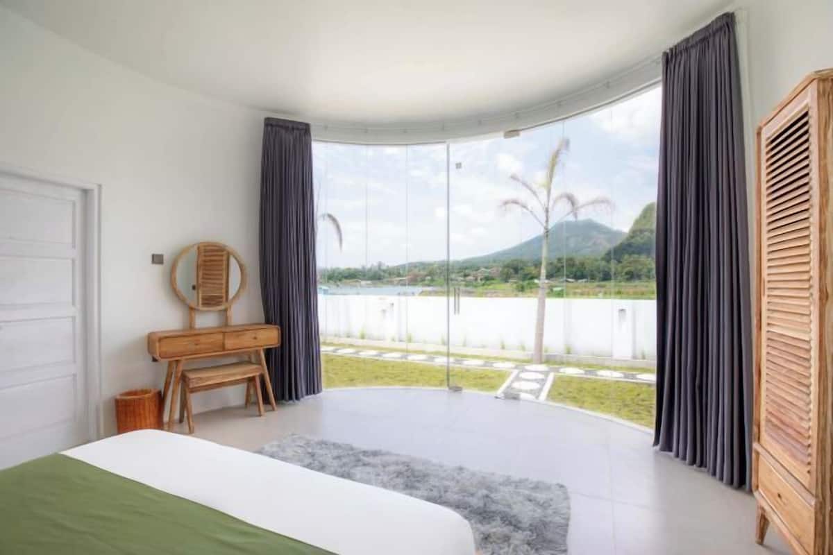 A bedroom with a bed, a wooden dresser, a mirror, and a view of a lake and mountains through large windows.