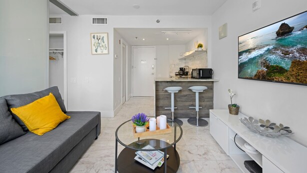 A modern living room with a grey sofa, a glass coffee table, and a kitchen area in the background. - Beachside Charm: Walk to the Beach (Miami Beach)