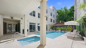 A swimming pool area with a glass door entrance, lounge chairs, and a white umbrella. - Beachside Charm: Walk to the Beach (Miami Beach)