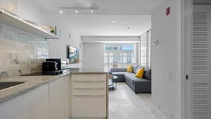 A modern kitchen with a built-in oven, a coffee machine, and a toaster. There is a grey sofa with yellow cushions in the living area, and a window with a view of buildings. - Beachside Charm: Walk to the Beach (Miami Beach)