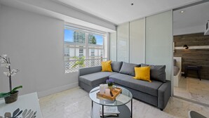 A modern living room with a grey sectional sofa, yellow cushions, a glass coffee table, and a view of buildings through large windows. - Beachside Charm: Walk to the Beach (Miami Beach)