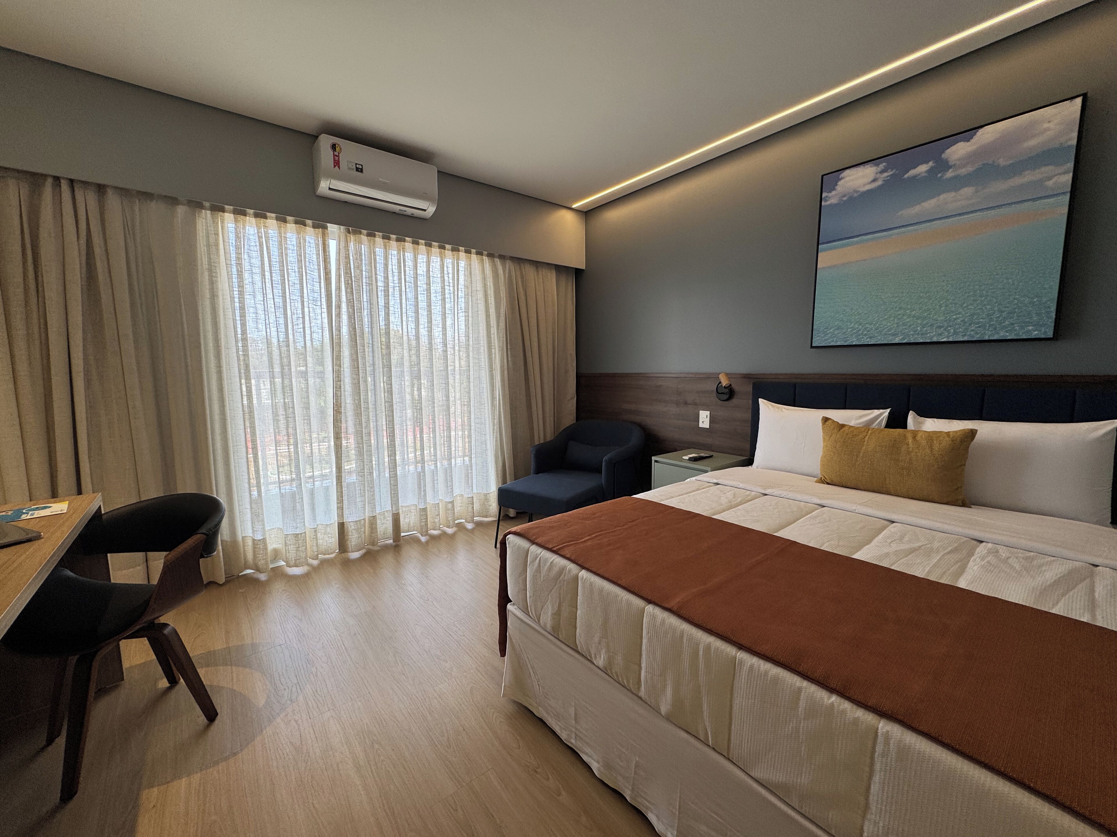Una habitación de hotel con cama, escritorio, silla, mesita de noche y un cuadro en la pared.