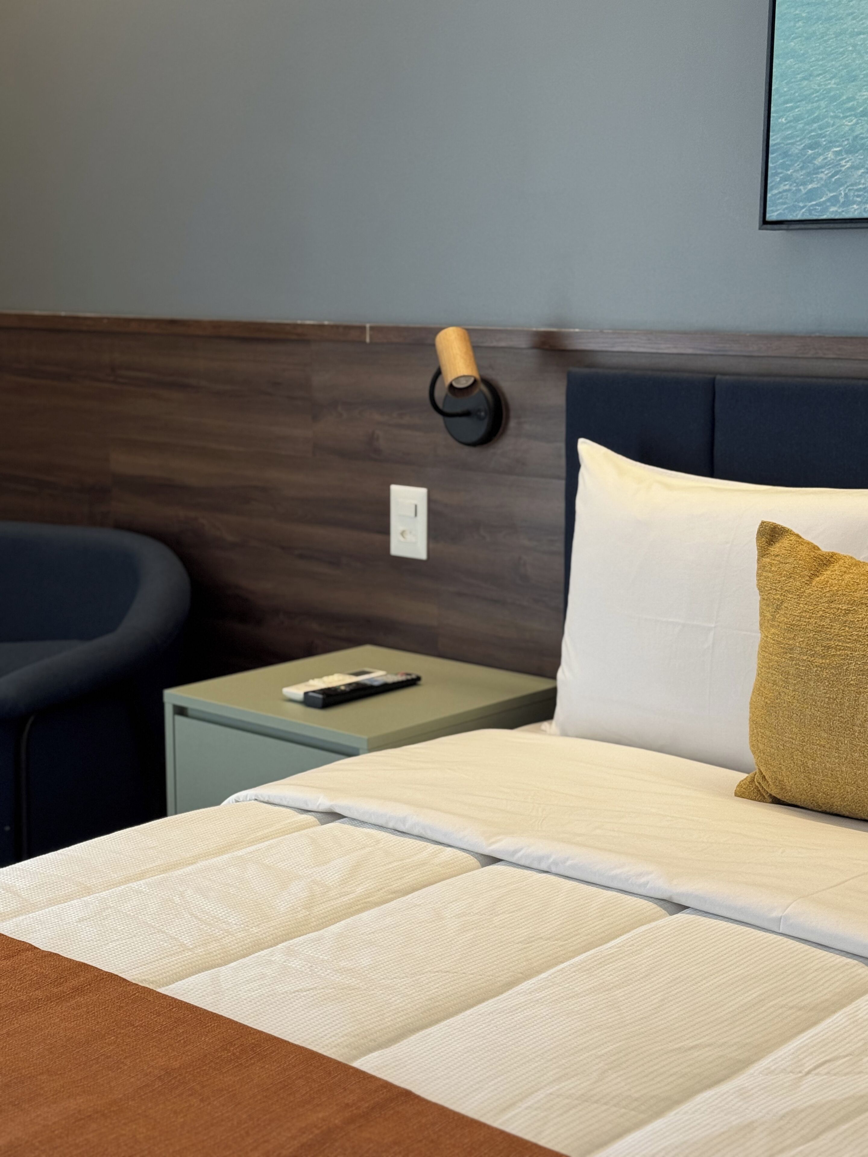 A neatly made bed with a white and gold pillow, a bedside table with a remote control, and a wall-mounted lamp.